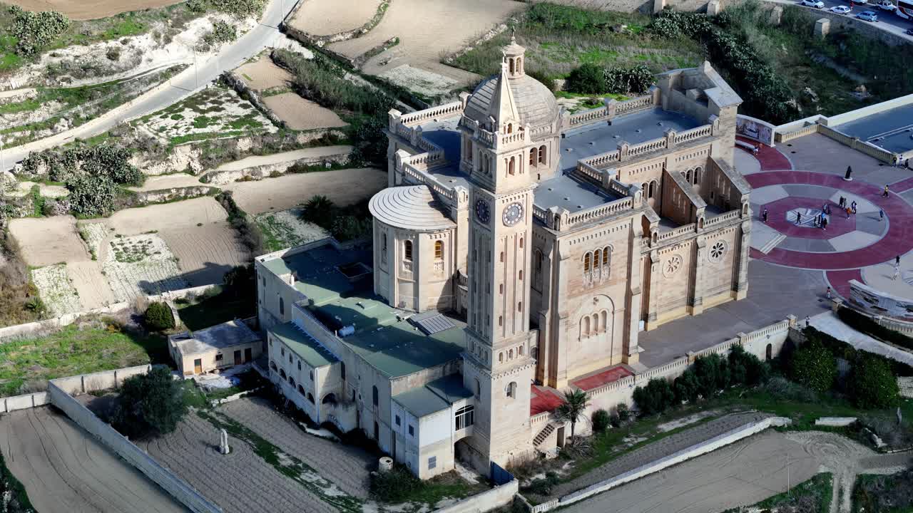 Veduta aerea della maestosa Basilica di Ta' Pinu a Gozo, Malta, durante una luminosa e soleggiata mattina d'inverno, con architettura in pietra calcarea dorata su campi terrazzati verdi sotto un cielo azzurro e limpido.