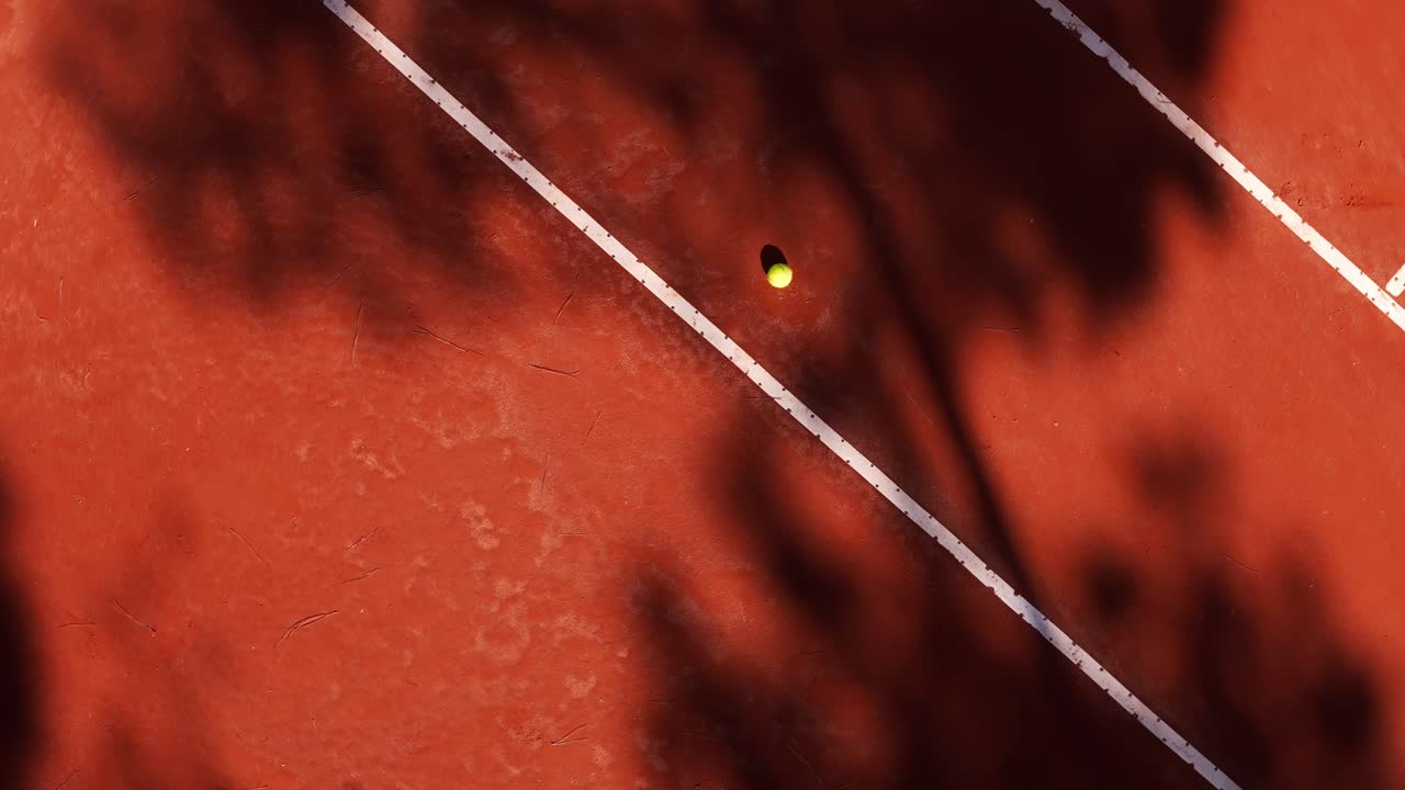 Rotating aerial drone shot revealing a tennis ball and tree shadows on a clay court in Italy, Italia, creating a dynamic contrasting scene