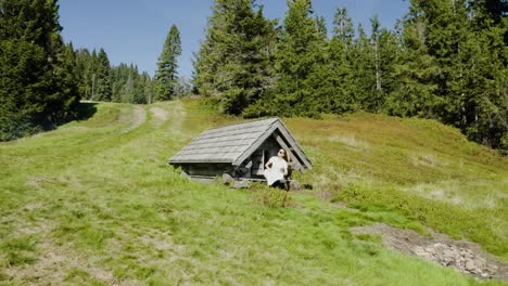 young woman reads unfolded map, looks for a trail, sitting in front of small old wooden mountain hut, placed in the middle of mountain clearing