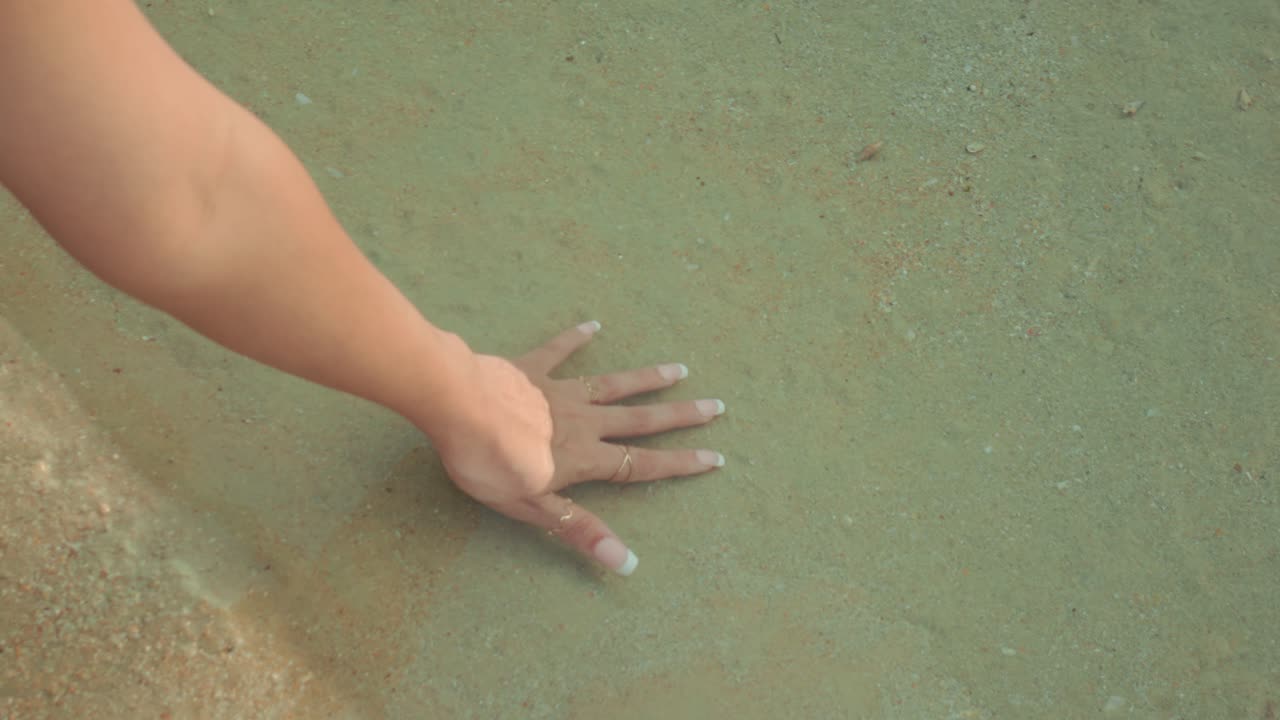 Manicured female hand resting in calm shallow beach water under soft light