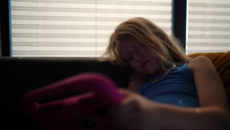 young girl experiencing boredom, yawning while using pink tablet and sitting comfortably on sofa near window with closed blinds, embodying modern digital lifestyle