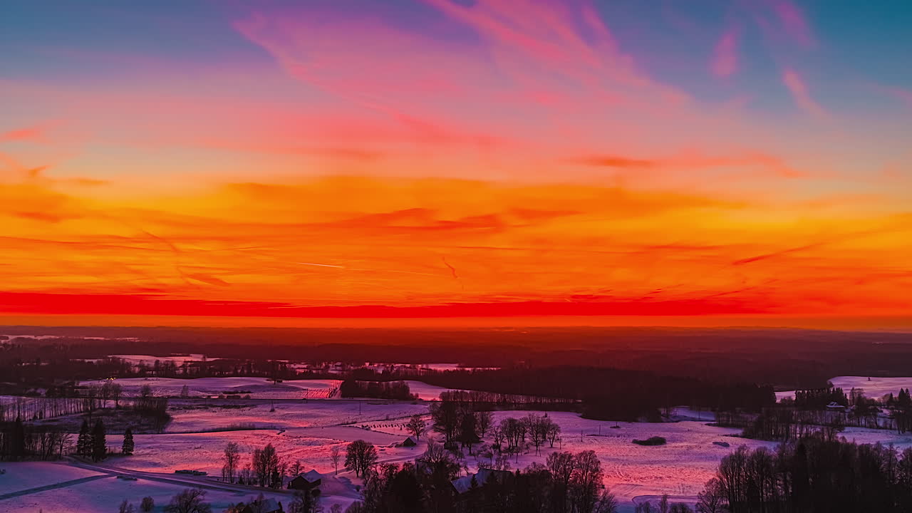 Winter sunset with dramatic orange sky hues over the vast snowy fields and distant forests in drone hyperlapse