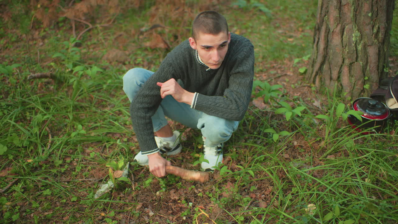 Man in sweater crouches on forest ground using wooden stick to fix peg into soil with yellow rope, surrounded by grass and trees, calm atmosphere with cooking plate and bag nearby