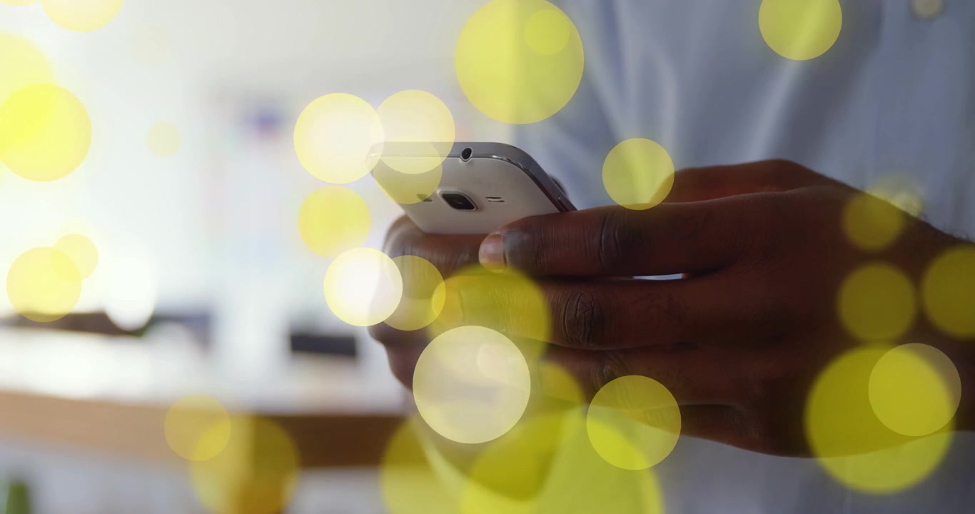 Homme tenant un smartphone blanc, naviguant dans une application en tapotant avec le pouce, avec une superposition de bokeh jaune masquant.