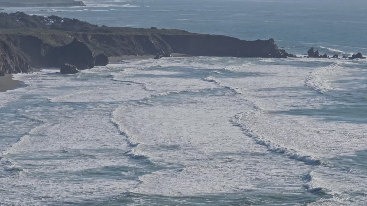 A wide, sweeping drone shot captures foamy waves rhythmically crashing onto the rugged shores of California’s dramatic coastline near Big Sur.