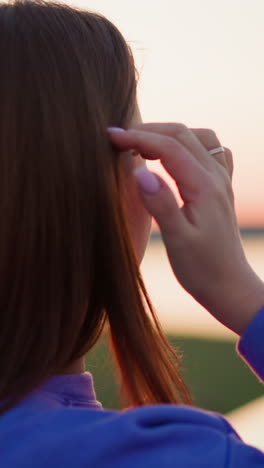 woman puts hair behind ear in dusk park closeup. tranquil lady adjusts hairstyle walking in twilight meadow. female walker in spring nature