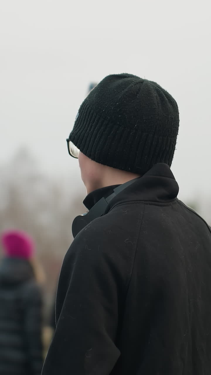 primer plano de un individuo con un traje negro y gorra, mirando hacia un campo, el fondo presenta una vista borrosa de personas que pasan