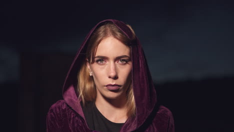 mysterious young blonde woman wearing a hood and a cloak and staring at the camera with serious expression