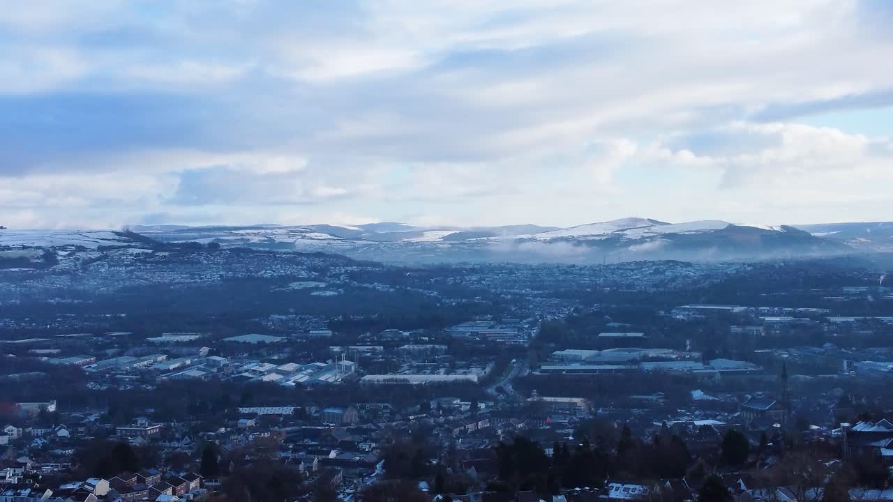 Luftfoto af sneklædte bakker omkring Swansea bymiljø med lavthængende skyer i dalen med en blanding af huse, veje og industriområder nedenfor. Snapshot af vintermiljøet i Wales.