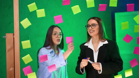 ladies discuss the ideas written on the sticky notes. women take the notes from glass wall and throw them back.