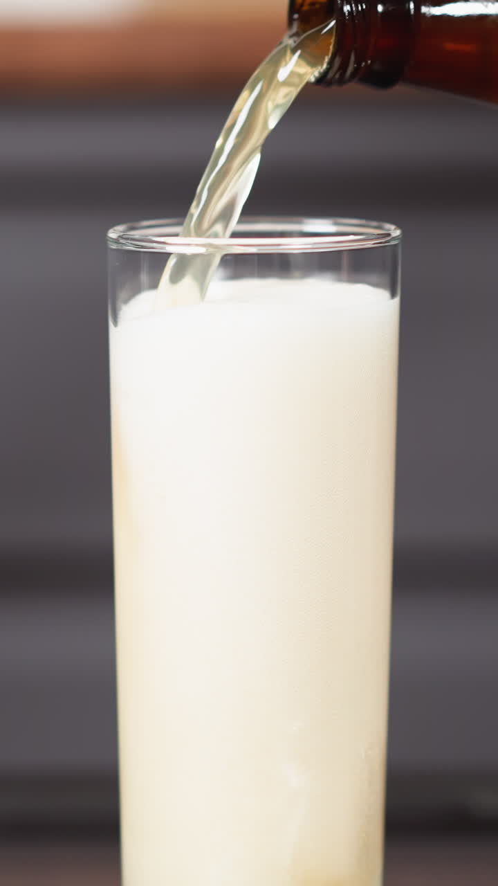 verter una bebida cremosa en un vaso alto, vista parcial de la mano con la botella, superficie reflejada