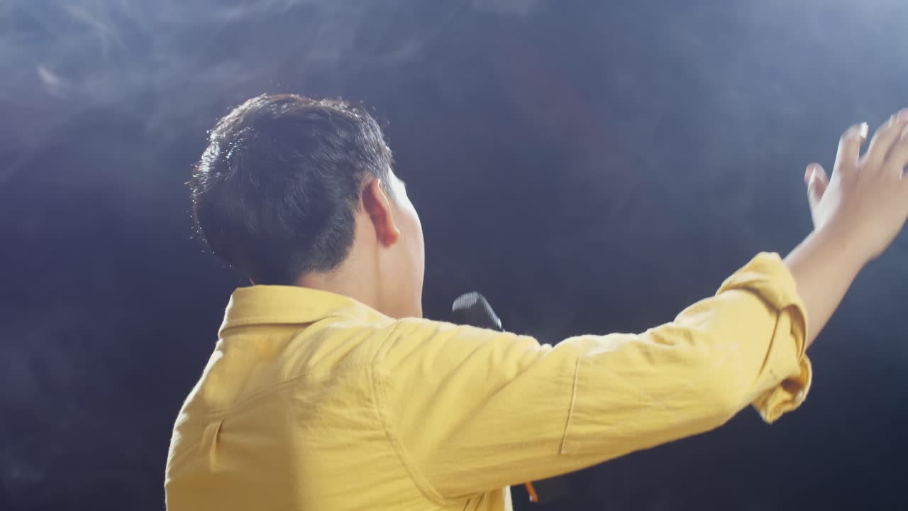 젊은 아시아 소년이 마이크를 들고 색 연기와 검은색 배경에 을 치는 가까운 뒤의 모습