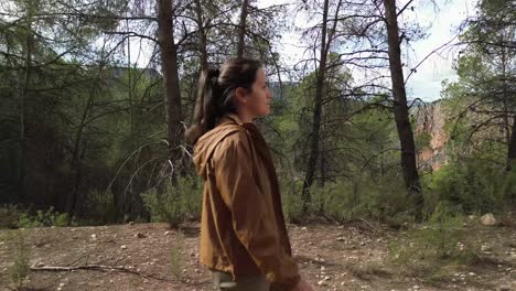 side tracking shot of a happy, attractive girl in a brown jacket walking through a pine and rosemary forest with a mountain in the background, enjoying nature