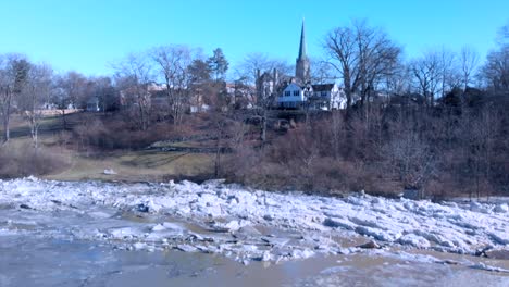 drone-shot-pan-up-shot-flying-over-partially-frozen-and-cracked-river-to-houses,-bare-trees,-and-church-steeple