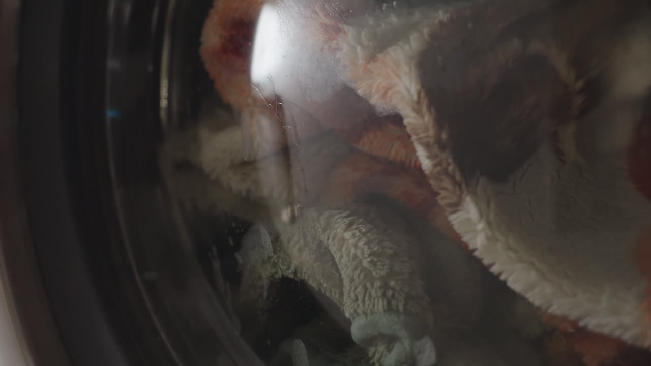 Zoom in on stainless washing machine as drum spins behind curved glass then pauses; rim reflections and soft blur highlight motion to stillness, soap traces on window, metallic laundromat mood
