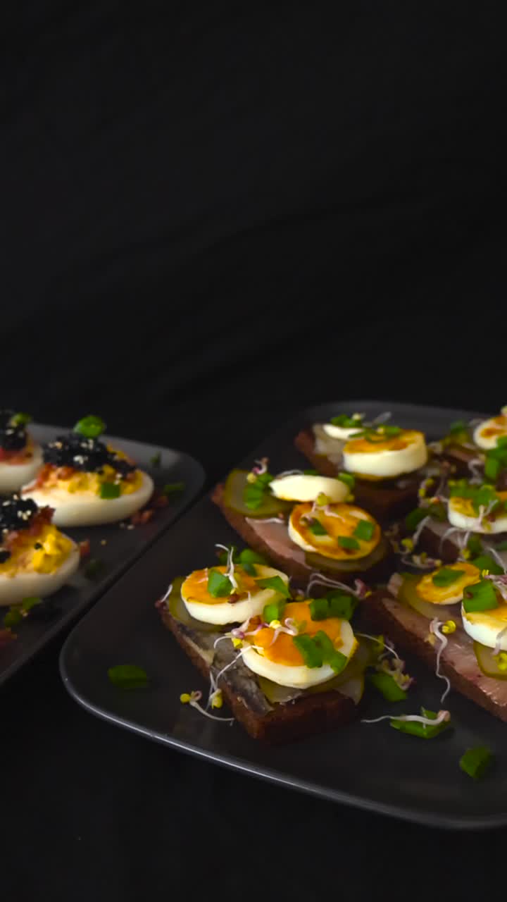 To elegante tallerkener med lækre forretter, den ene med fyldte æg toppet med sort kaviar og bacon, og den anden med brislingemadder på mørkt brød med syltede agurker og spirer.