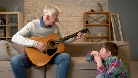 grandpa plays for his grandson on guitar, music. a young fat guy is sitting on the haven with his grandfather. teaches his grandson playing the guitar. home comfort, family idyll, cosiness concept, difference of generations 60 fps