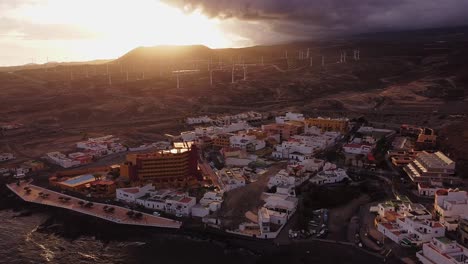 Impresionante-Vista-Aérea,-Ciudad-Con-Casas-Y-Molinos-De-Viento-En-La-Playa-Costera-De-La-Isla-De-Destino-Turístico-Tenerife-Durante-La-Hora-Dorada-Del-Sol-De-Verano,-Panorámica