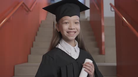 zoom in of a happy asian preschool female student in cap and gown holding graduation diploma and looking at the camera