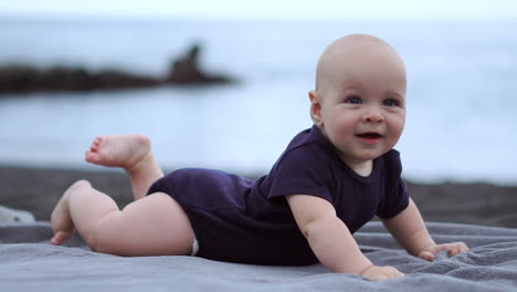 Nestled-on-the-black-sand-near-the-ocean,-the-baby-lies-on-his-stomach,-erupting-in-laughter-while-engaging-with-the-camera