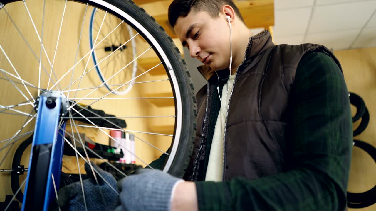Concentrated mechanic is looking at rotating bicycle wheel checking spokes then straightening them with tools. Young man is wearing greasy gloves and listening to music.