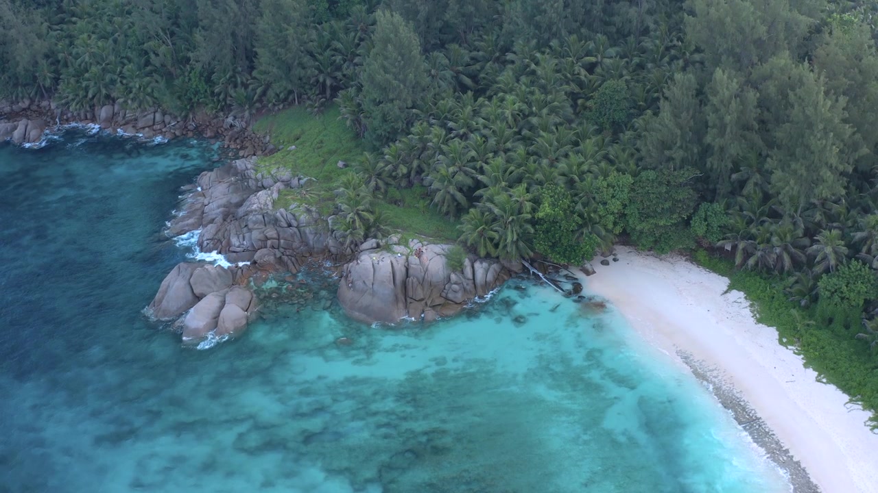 Ripresa aerea con drone che panoramica su uno spettacolare promontorio granitico delle Seychelles con massi, coperto da palme da cocco, con una piccola insenatura riparata di acqua turchese brillante e una stretta spiaggia sabbiosa sottostante