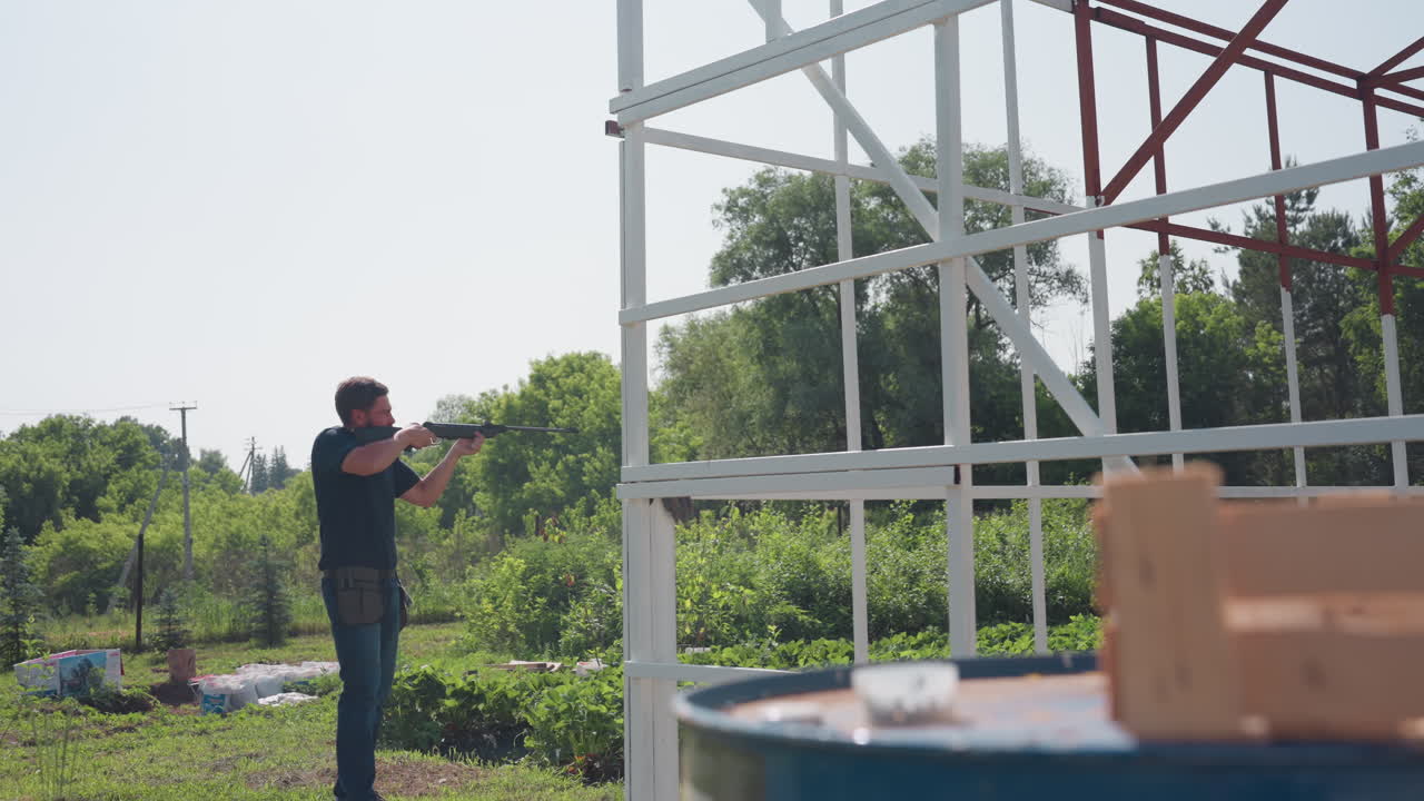 man uses rifle to scare birds near drum close to crate and container on farm frame while inspecting crops under bright sun, tense protective gesture keeping wildlife away from plants