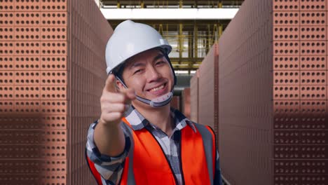 close up of asian male engineer with safety helmet smiling and touching his chest then pointing at you while standing with red brick packed in stacks are stored