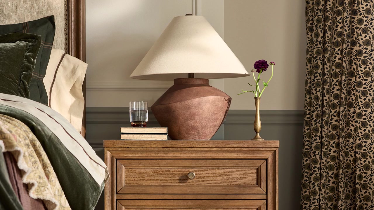Slow zoom out of a farmhouse-style table lamp on a rustic nightstand with flowers and a glass of water in a professionally staged bedroom.