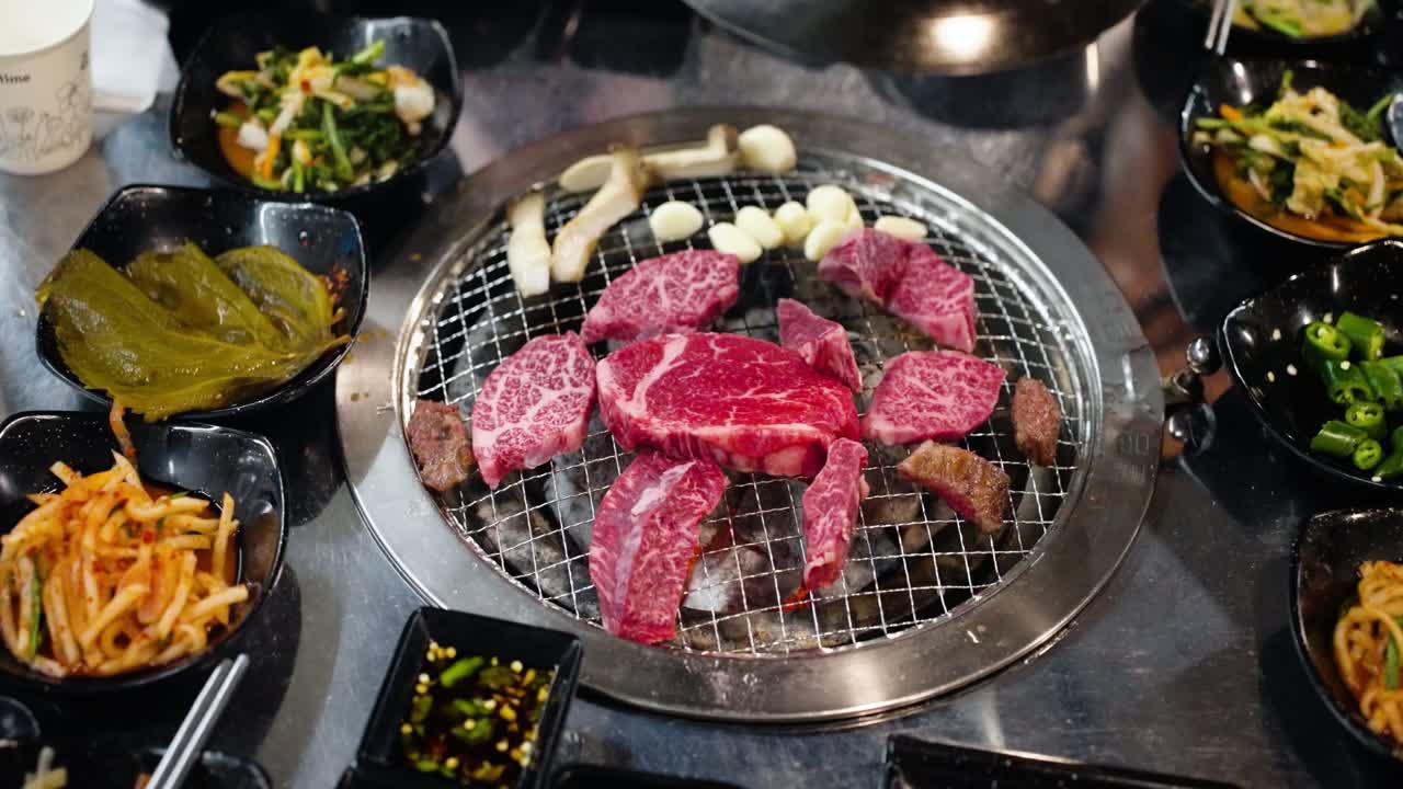 Top-down view of beautifully marbled raw Hanu beef slices, mushrooms, and garlic cooking on a circular charcoal grill, surrounded by an assortment of delicious Korean banchan side dishes