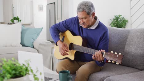 Video-of-happy-senior-biracial-man-using-laptop-and-playing-acoustic-guitar-at-home