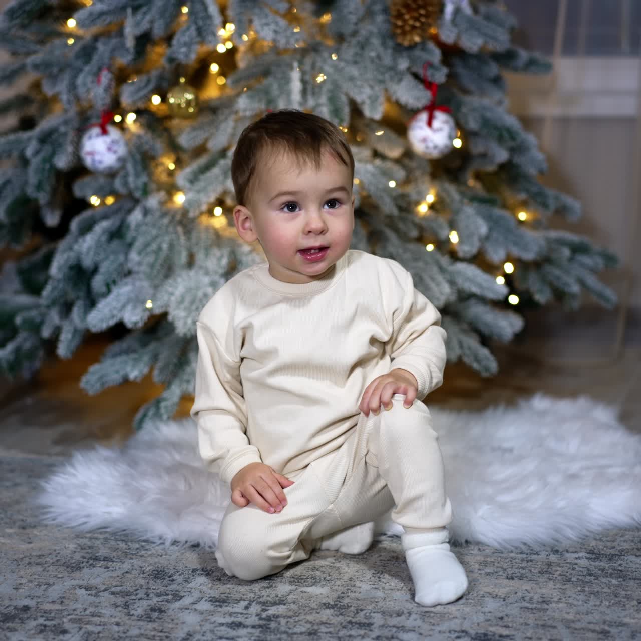 Little cute angelic baby sitting near Christmas tree in the room. Lovely kid sits still as if waiting for something and then smiles adorably as camera approaches
