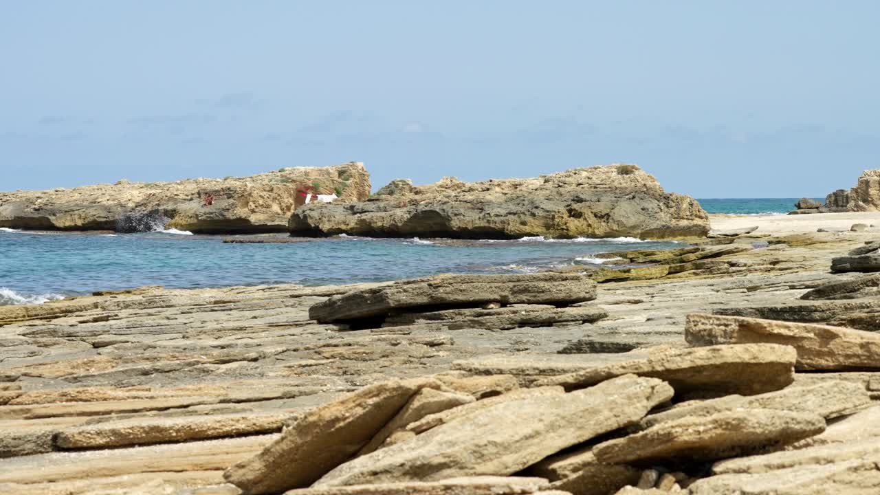 Waves crash gently against unique layered sandstone rocks on a rugged coastline under clear blue sky and bright sunlight.