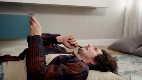 A-thoughtful-guy-comes-with-curly-hair-lying-on-a-pillow-and-writing-down-his-ideas-in-a-notepad-while-lying-on-the-floor-in-a-modern-apartment