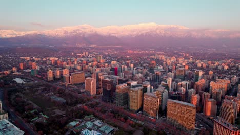 Paralaje-Aéreo-De-Los-Edificios-Del-Distrito-Financiero-De-Las-Condes,-Montañas-Cubiertas-De-Nieve-En-El-Fondo-Al-Atardecer,-Santiago,-Chile