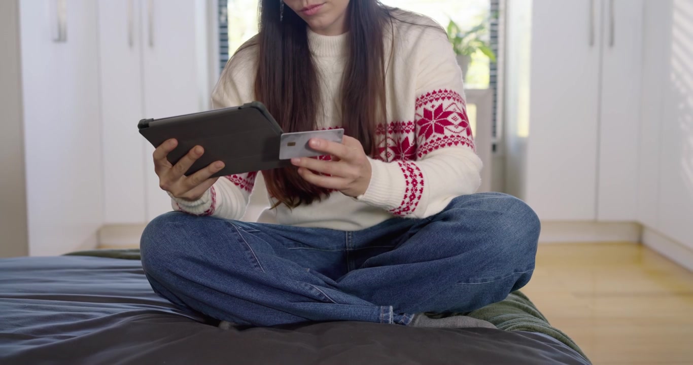 Adult female in knit sweater and jeans scrolling tablet on bed, picking card for verification