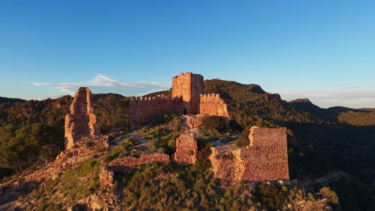 Rovine di un castello medievale in cima a una collina rocciosa nella Sierra Calderona, Spagna, bagnate da una calda luce dorata con montagne boscose e cielo blu sullo sfondo.