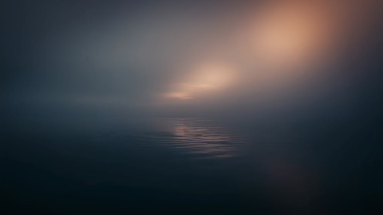 Brightening diffuse warm glow shifting up over calm water at dawn through fog, reflecting ripples