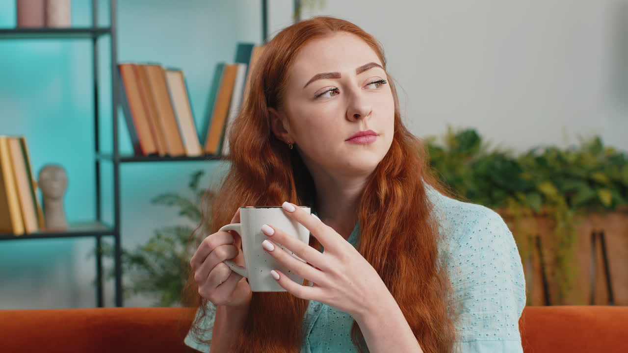 Attractive smiling young redhead woman drinking a cup of coffee or herbal tea at home couch