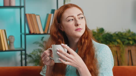 attractive smiling young redhead woman drinking a cup of coffee or herbal tea at home couch