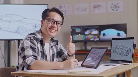 vista lateral de un hombre asiático sonriendo y mostrando el pulgar hacia arriba mientras dibuja el concepto de diseño de un nuevo automóvil en una tableta en el estudio con televisión y computadoras muestra modelo de automóvil eléctrico 3d