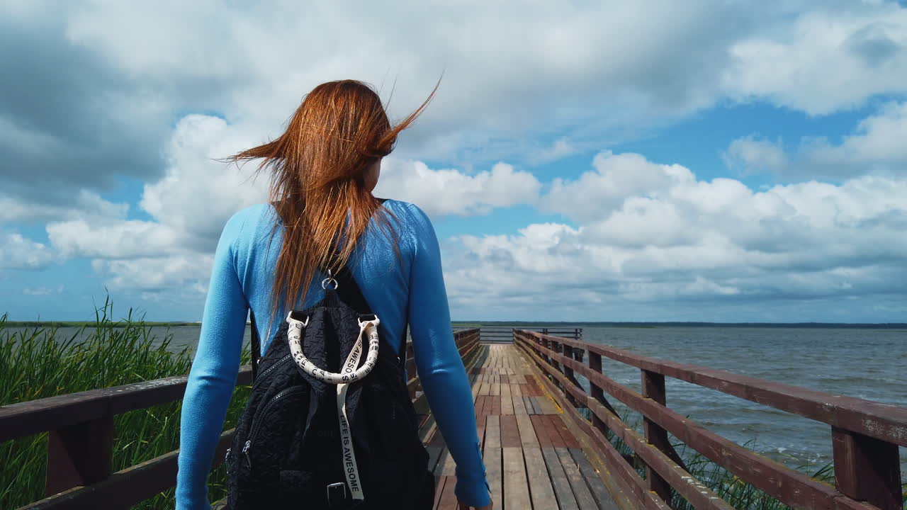 Giovane donna vista da dietro con i capelli al vento che cammina su un molo di legno su un lago