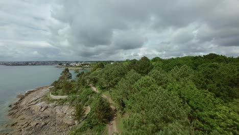 FPV drone pushes in and ascends above lush green trees in Concarneau, stunning view of sea beside them under dramatic cloudy sky in Brittany, France, beauty of coastal landscape.