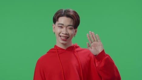 a smiling asian transgender male waving hand and say hi while standing on green screen in the studio