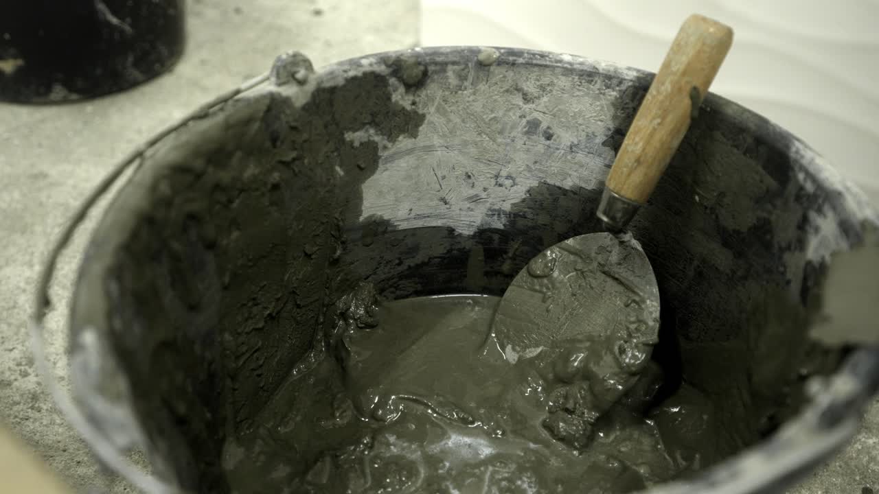 Bucket half full of cement with trowel, hand putting back drywall knife