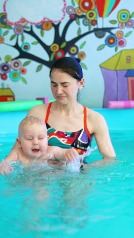 caucasian woman is holding a little blond baby boy in the swimming pool. kid waves his hands splashing water to the sides. vertical video.