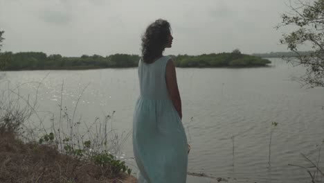 Static-slow-motion-cinematic-shot-of-a-young-and-pretty-model-in-turquoise-dress-with-wooden-basket-in-her-hand-looking-pensively-over-a-lake-with-sea-islands