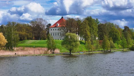biržai castle in mid-spring: a mid-angle view of the white-walled, red-roofed historic building surrounded by vibrant green hills and budding trees under a dramatic, partly cloudy sky