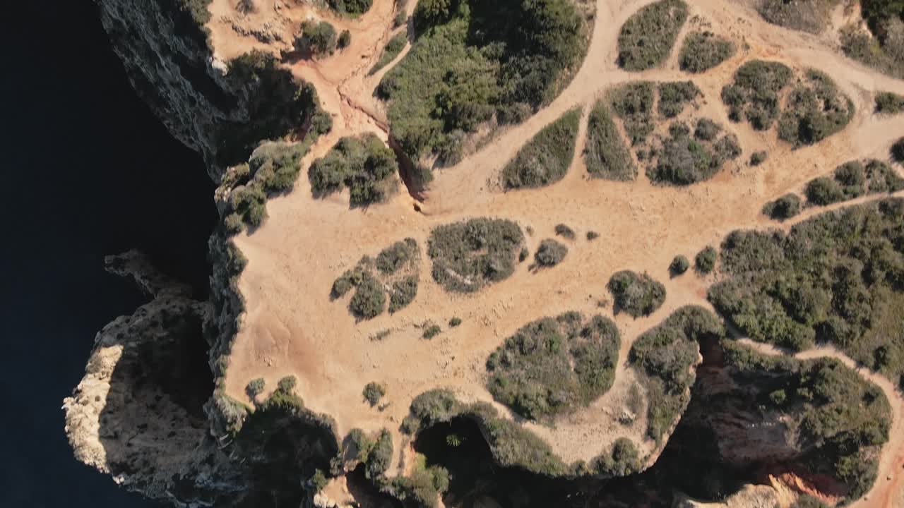 Overhead view from the drone reveals the intricate maze of Algarve’s limestone cliffs and sea arches, with patches of green shrubbery and hidden coves below, as the drone advances