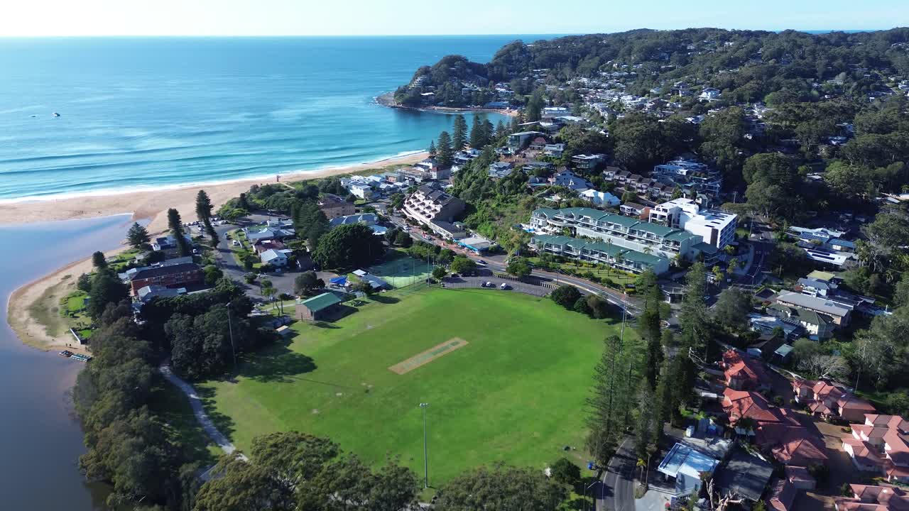 Drone aerial of Avoca Beach public sports oval Heazlett Park surrounded by residential housing town shops and tree-lined streets with ocean coastal waves breaking in background Central Coast Australia